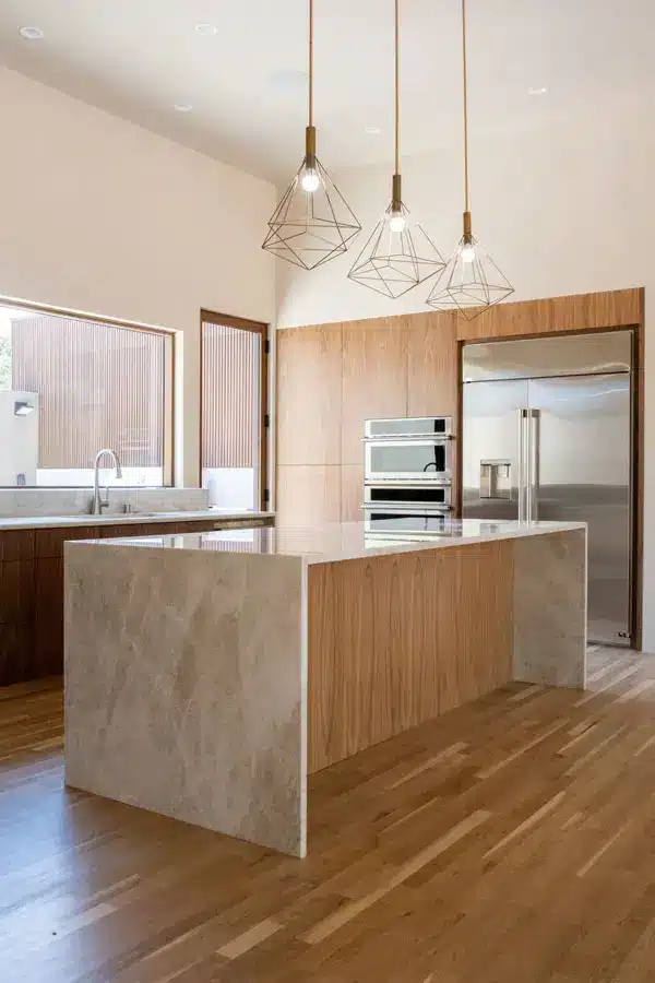 Modern kitchen with a light wood floor and a central island featuring a beige marble countertop. The kitchen has built-in stainless steel appliances, including a double oven and a large refrigerator. Three geometric pendant lights hang above the island.