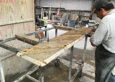 A worker polishes the edge of a large granite countertop on a metal cart in a workshop. Various tools and equipment are visible in the background. The countertop has a mottled brown and beige pattern.