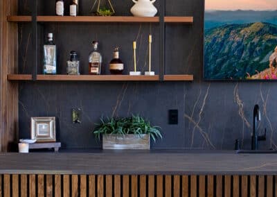 A modern home bar featuring dark stone backsplash, wooden shelves with decorative items, and a counter with vertical wood slats. Two white cushioned stools sit at the bar. Copper pendant lights hang above, and a landscape photo on the wall completes the scene.