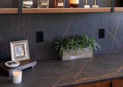 A home bar with dark marble countertops features several liquor bottles, a framed picture, a small plant in a rectangular planter, and a lit candle. The wall has two electrical outlets and is styled with dark marbled panels.