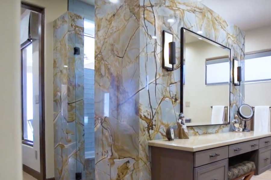 A bathroom features a large mirror above a double sink vanity with brown cabinets. The shower has a glass door and a striking marble wall with blue, gold, and white veins. A window allows natural light into the space.