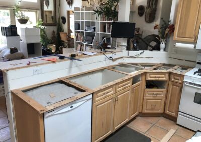 A kitchen undergoing renovation with light wood cabinets and no countertops. A dishwasher and stove are visible. Boxes and household items are scattered around. Walls have shelves and masks as decor.