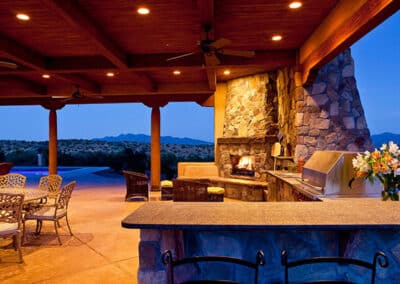 Outdoor patio with stone fireplace, grill, and bar counter. Wrought iron chairs and a table are on the terrace. The sky is clear, with hills visible in the background. A vase of flowers is on the counter, and the area is lit by recessed lights.