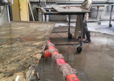 A worker in a cap stands at a table, crafting a large stone slab in a workshop. The floor is wet, and various tools and machinery are visible. A partially finished countertop is in the foreground.