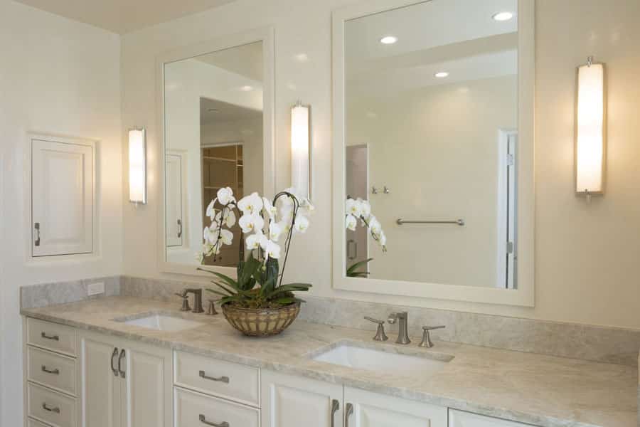A bathroom with a double sink vanity, light gray countertop, and two large mirrors. Each sink has a modern faucet, and there is a potted orchid plant between them. Overhead lights and wall sconces illuminate the space. White cabinets provide storage.
