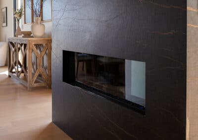 A modern indoor fireplace set in a sleek, dark marble wall. The room features light wood flooring and a wooden console table with decorative vases and artwork in the background.