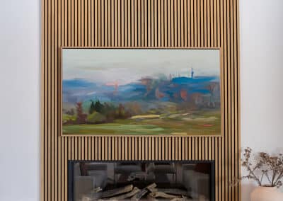 A modern fireplace set in a wall with vertical wooden slats. Above the fireplace, there's a landscape painting featuring rolling hills and a distant horizon. A white vase with dried branches is placed on the ledge to the right.