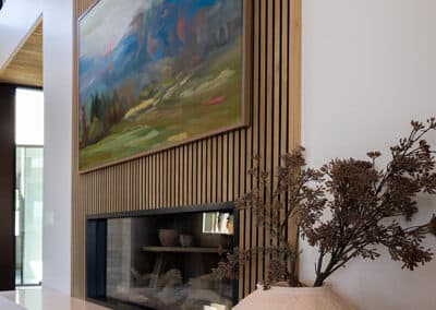 A modern interior with a large landscape painting on a wooden slat wall. Below is a black-framed fireplace with a white marble mantel. A white vase with dried branches is placed on the mantel, adding a natural touch to the decor.