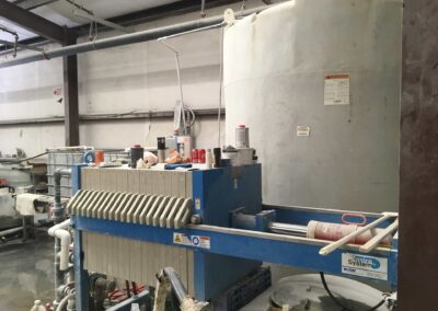 Industrial machinery inside a factory setting, featuring a large cylindrical tank and a blue filtration unit. The area is cluttered with various tools and containers. Overhead pipes and metal beams are visible as part of the infrastructure.