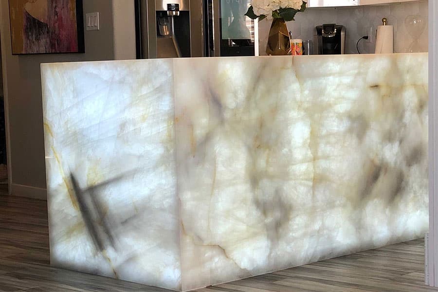 A kitchen island made of translucent stone is lit from within, showcasing its marbled texture. The surrounding kitchen includes modern appliances and decor elements like a vase with flowers on the counter.
