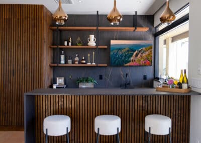 A modern home bar featuring wood paneling and three white stools. Shelves hold decorative items and bottles. Three copper pendant lights hang from the ceiling. A landscape photo is mounted on a dark textured wall. A wine tray is on the counter.