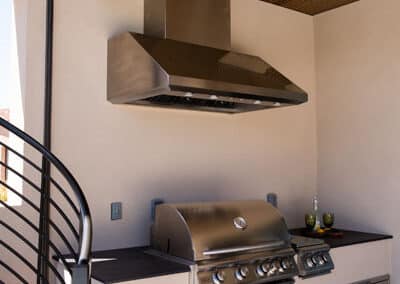A modern outdoor kitchen setup featuring a stainless steel grill with multiple burners, a large metallic range hood above, and built-in storage cabinets below. The area is covered by a wooden slatted ceiling, with a spiral staircase nearby.