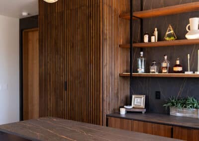 Modern kitchen with wooden slatted cabinetry and shelves. A dark stone countertop extends from an island, with two round stools in front. Two pendant lights with amber glass shades hang from the ceiling. A variety of bottles and decor items are on the shelves.
