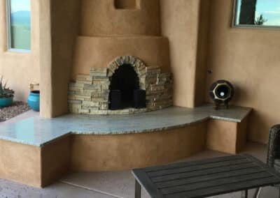 A stucco outdoor fireplace with a stone arch detail on the front and a wide stone bench. There is a decorative item on the bench to the right. Surrounding the area are a table and some potted plants.