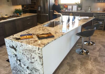 Modern kitchen with a large, marble island countertop featuring a sink and two bar stools. A person stands in the background near dark wood cabinets and stainless steel appliances. Brochures are displayed on the counter.