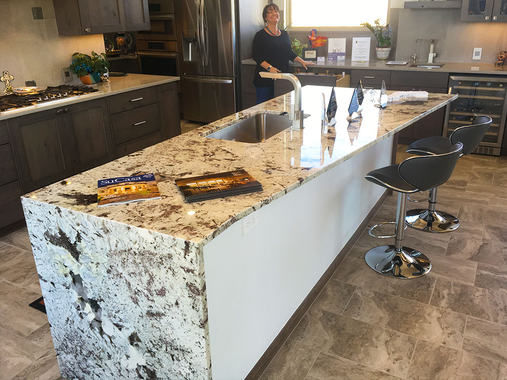 Modern kitchen with a large, marble island countertop featuring a sink and two bar stools. A person stands in the background near dark wood cabinets and stainless steel appliances. Brochures are displayed on the counter.