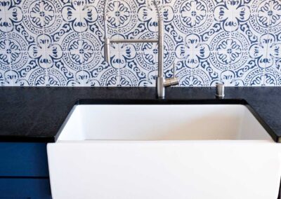 A kitchen sink with a white farmhouse-style basin is set against a patterned blue and white tile backsplash. The countertop is made of dark stone, and there is a silver faucet with a single handle on the right side.