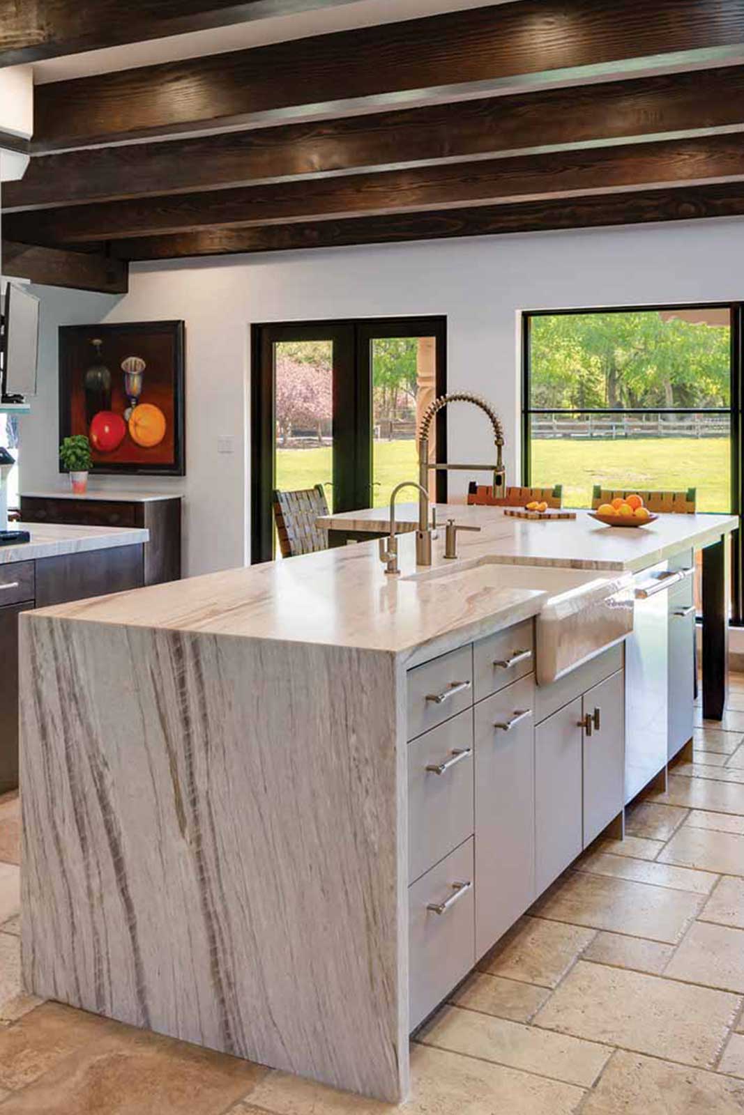United-Stoneworks-Kitchen-Countertops-Waterfall-White-Grey-Stripes A modern kitchen features a marble island with a built-in sink and faucet, white cabinetry, and a tiled floor. Large windows reveal a green outdoor view, and fruit bowls decorate the countertops.